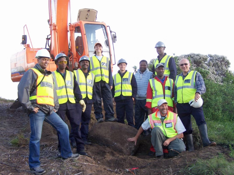 Professor Ellen Rathje smiling with construction crew at dig site in front of crane while wearing hard hat and safety gear